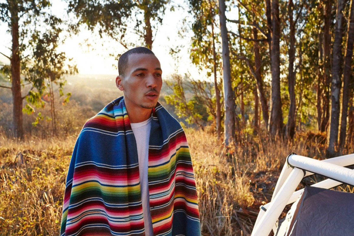 Man in blanket stands outside tent near forest. Soft sunset light illuminates the campsite in the outdoor adventure and travel image.