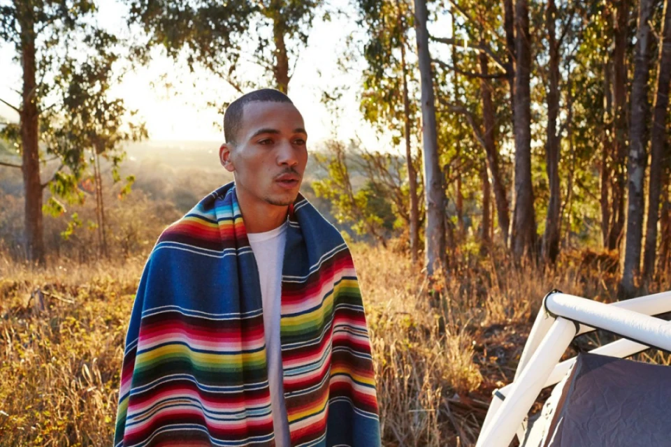 Man in blanket stands outside tent near forest. Soft sunset light illuminates the campsite in the outdoor adventure and travel image.