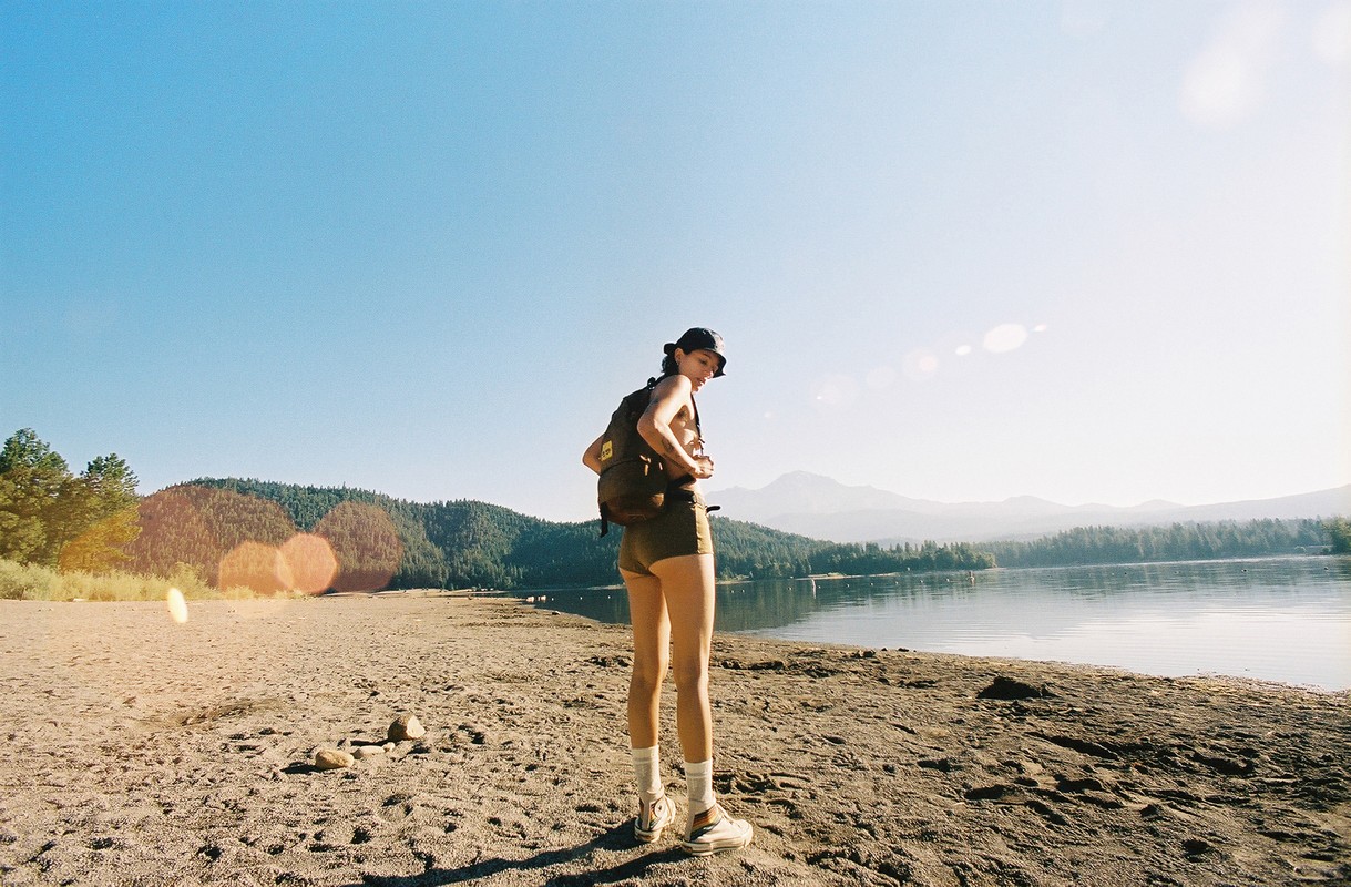 Woman with backpack hiking on sandy lake beach with mountains, blue sky, and sunshine; travel and nature photography in daylight.