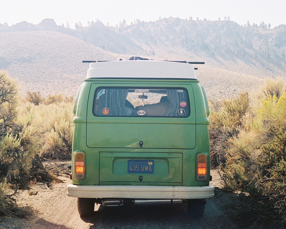 Rear view of a vintage green and white VW bus on a dirt road, surrounded by sagebrush, heading towards distant hills under a bright sky.