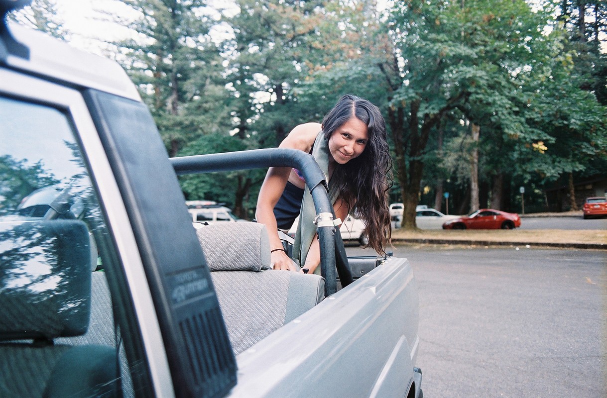 Woman leans into the bed of a pickup truck in an outdoor setting, with trees and other cars in the background. She smiles towards the camera.