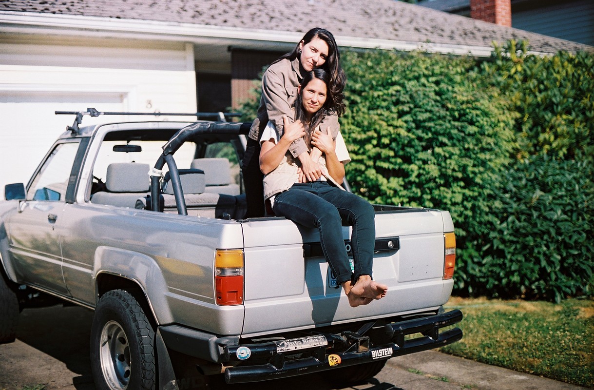 Two adult women sitting in the back of a vintage pickup truck in a residential area with green bushes and a house visible.