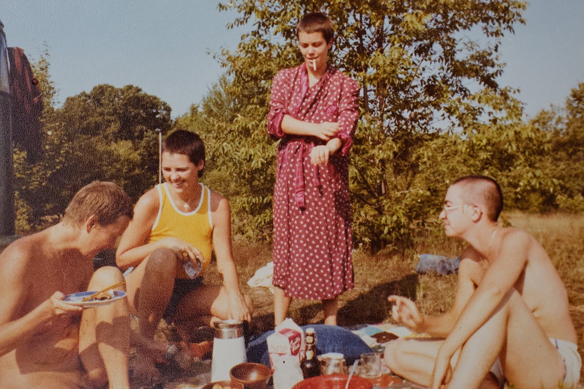 Outdoor picnic scene featuring four people; one woman in a floral robe smokes, and three others eat, set against a leafy backdrop.