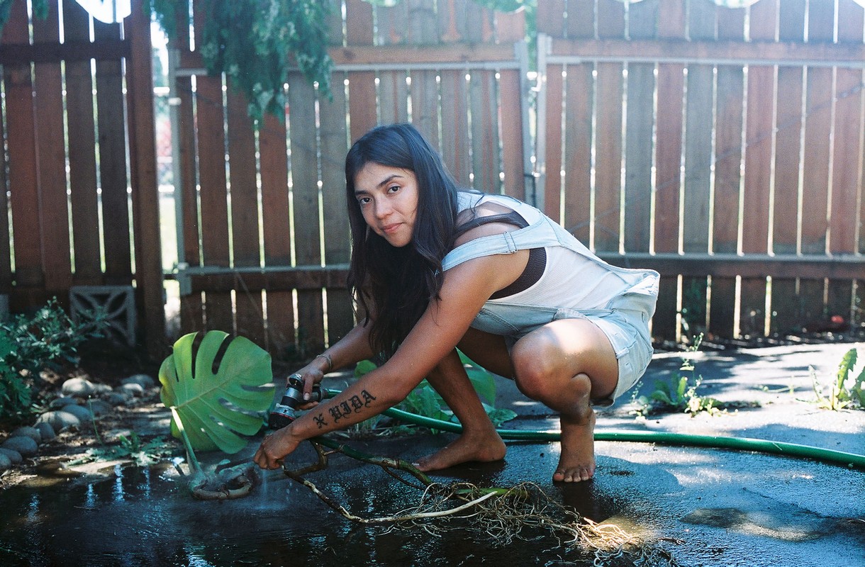 A woman in denim overalls crouches outside watering a monstera plant with a garden hose, with a weathered wooden fence in the background.