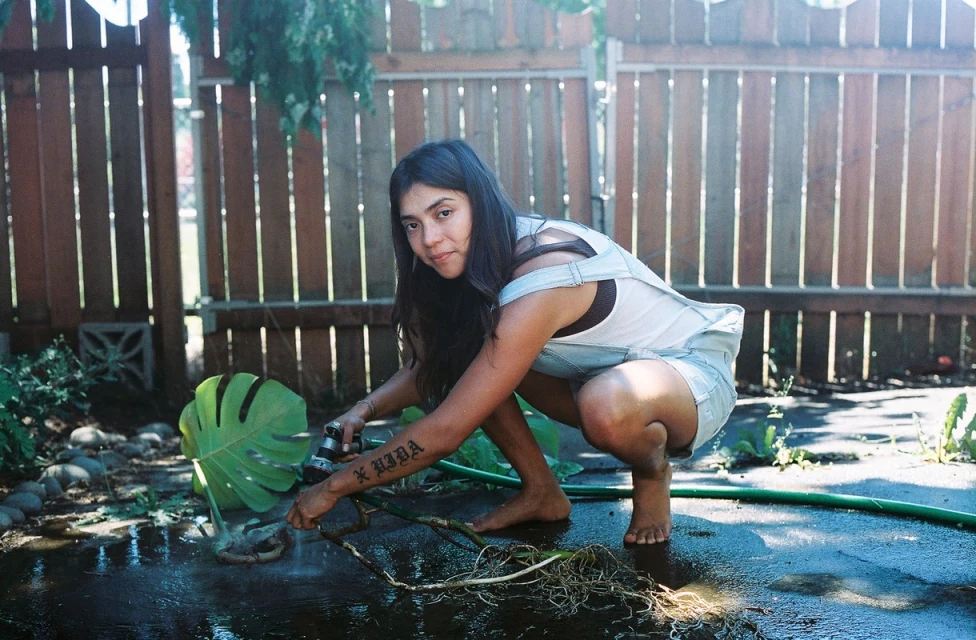 A woman in denim overalls crouches outside watering a monstera plant with a garden hose, with a weathered wooden fence in the background.