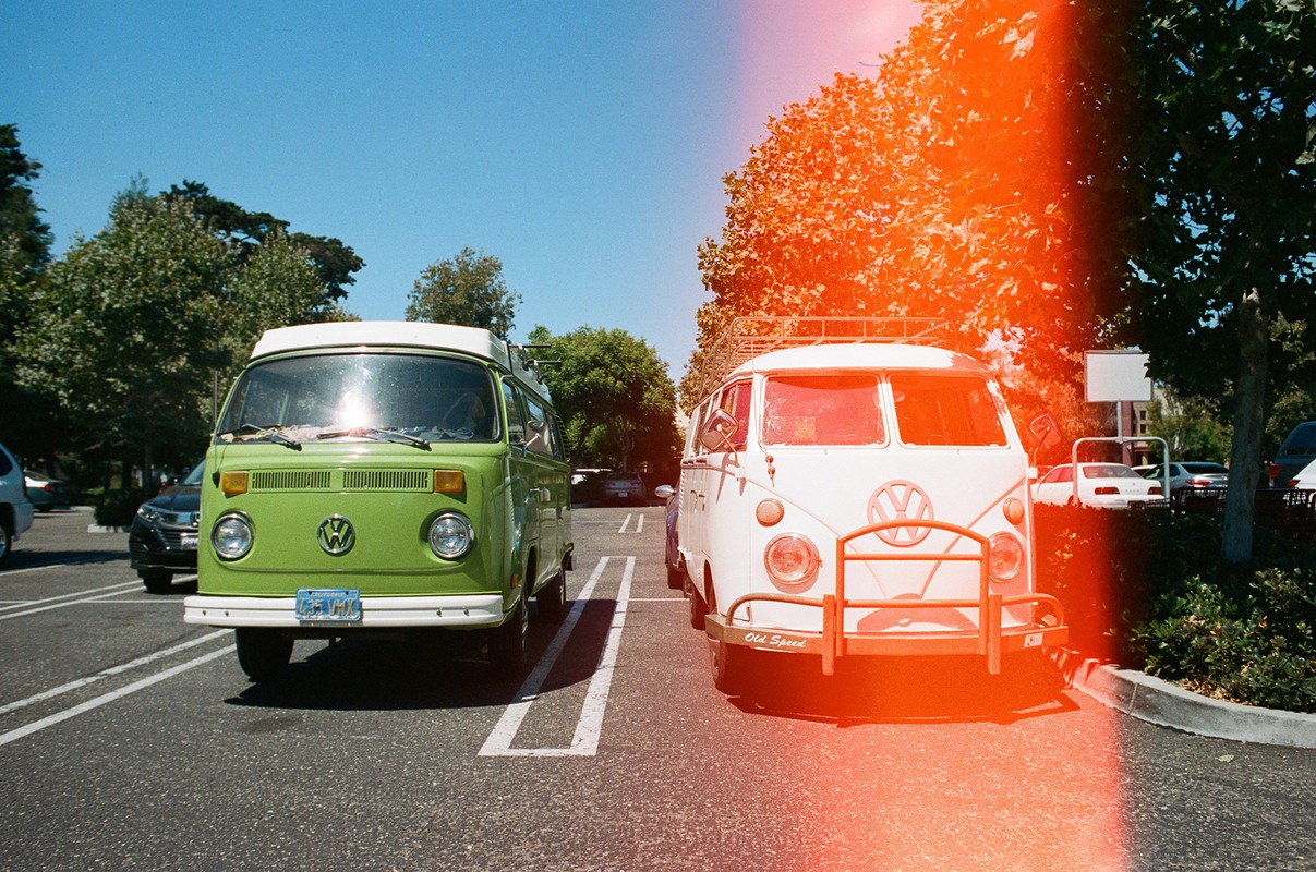 Two vintage Volkswagen vans parked in a row in a parking lot with trees and blue sky, creating a nostalgic and colorful scene.