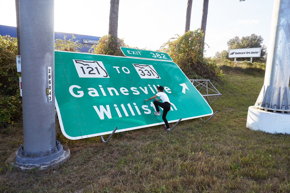 Young person climbs on a large, green highway sign for Gainesville and Williston in a grassy field; highway and tire center in background.