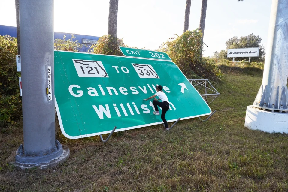 Young person climbs on a large, green highway sign for Gainesville and Williston in a grassy field; highway and tire center in background.