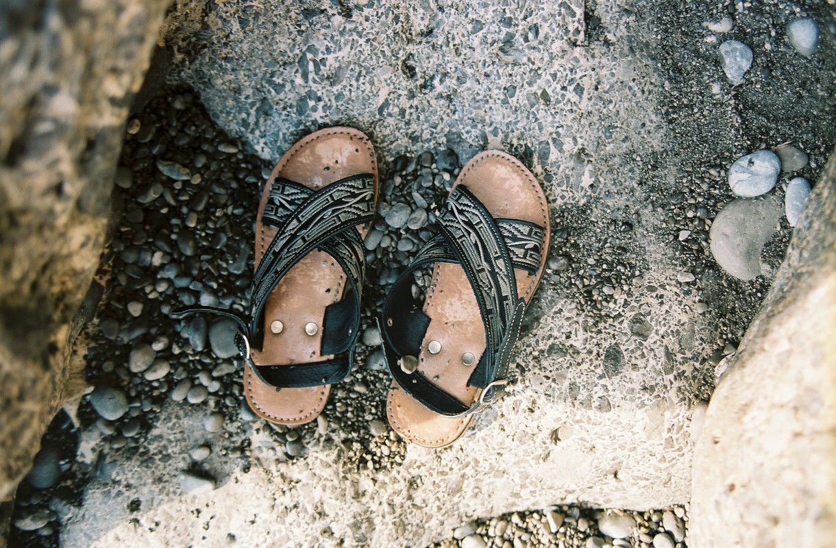 Overhead shot of well-worn leather sandals with patterned straps, resting on a textured surface with small pebbles in natural light.