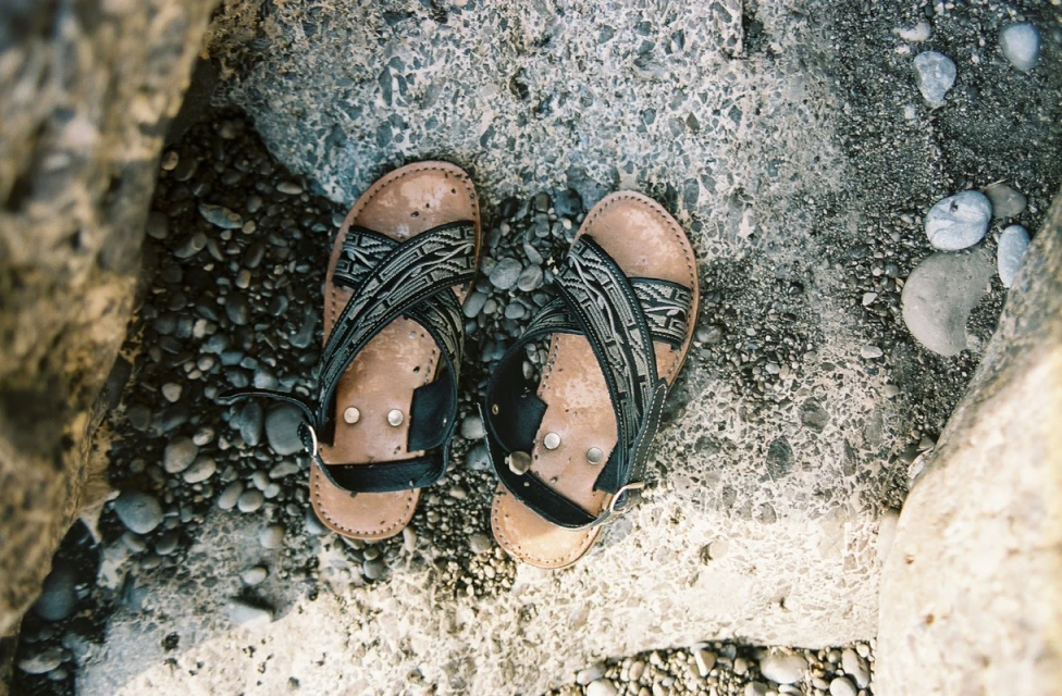 Overhead shot of well-worn leather sandals with patterned straps, resting on a textured surface with small pebbles in natural light.