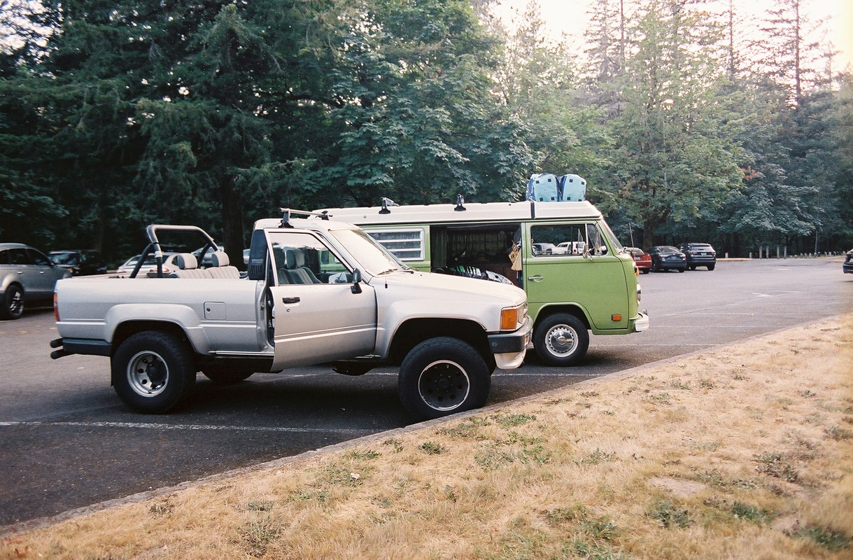 Silver pickup truck and green van parked side-by-side on asphalt lot near a forest, captured in an eye-level medium shot.