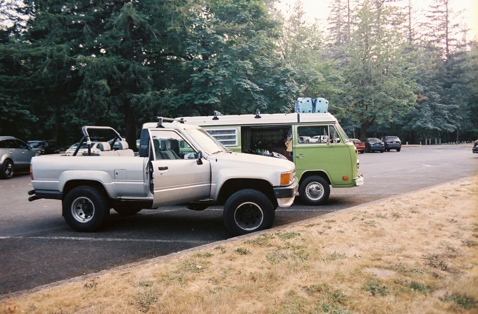 Silver pickup truck and green van parked side-by-side on asphalt lot near a forest, captured in an eye-level medium shot.
