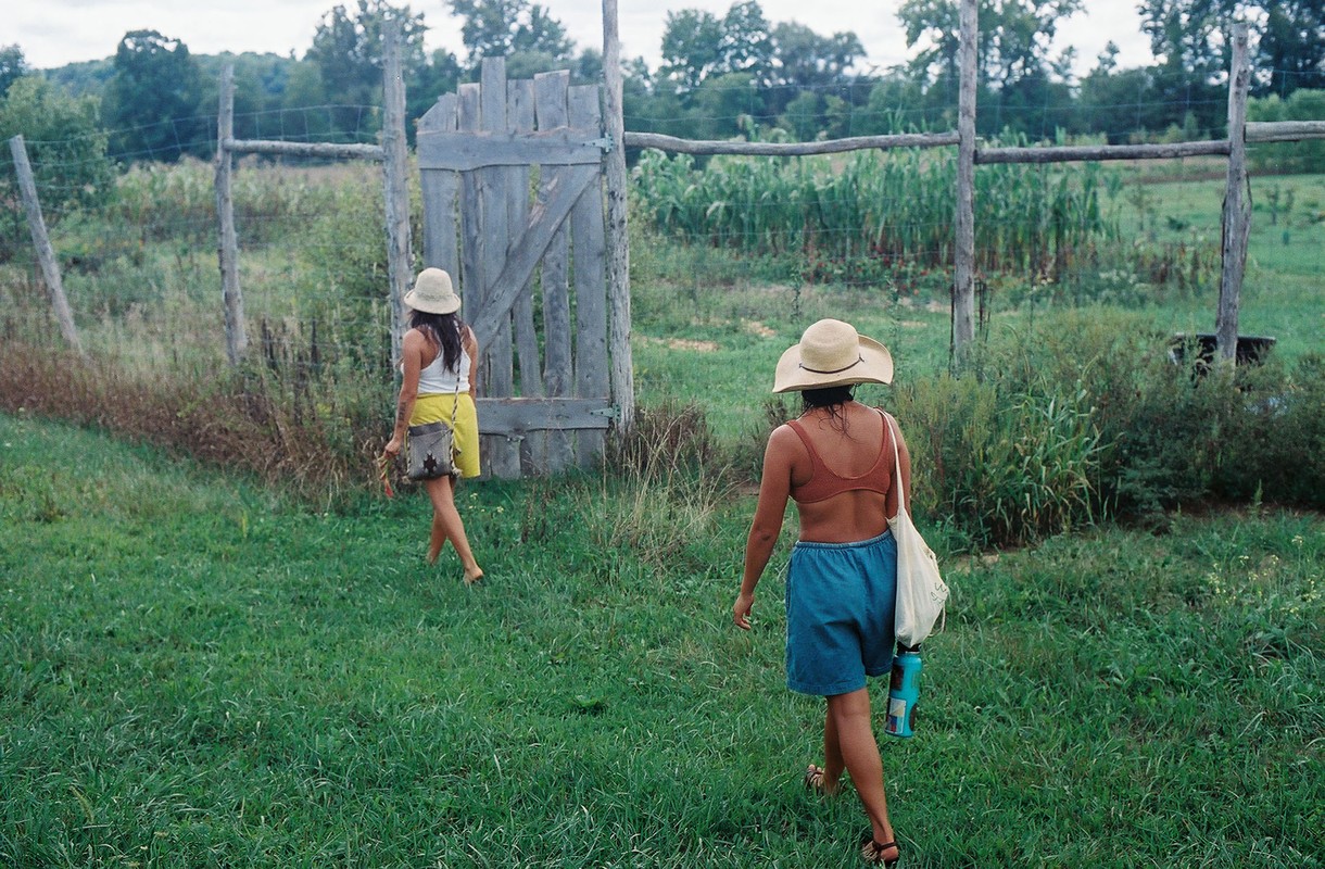 Two women in sunhats walk through a grassy field towards a wooden gate in the countryside on a sunny day, carrying bags.