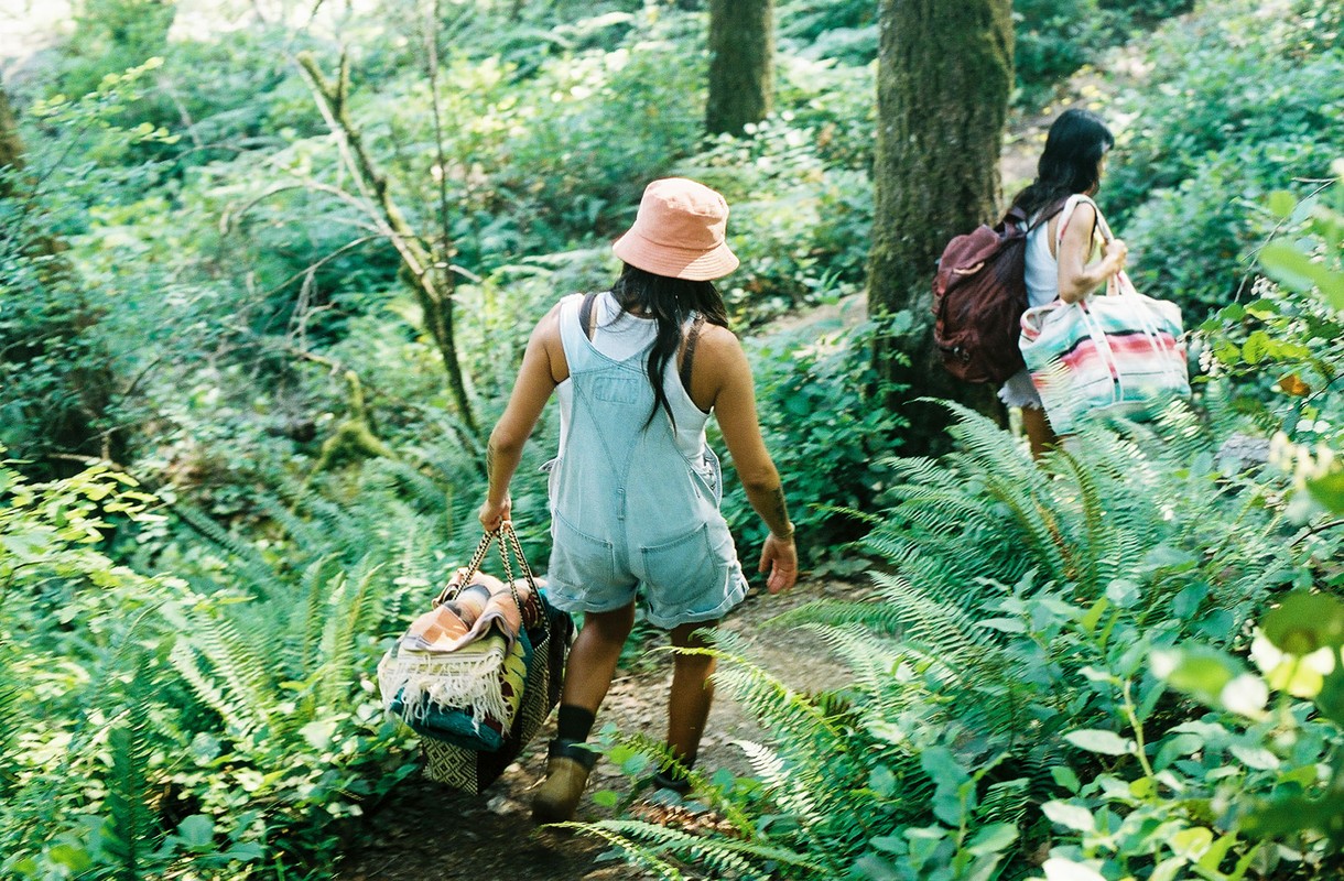 Two women hike through a lush green forest, carrying bags, and wearing casual summer clothes, captured in a scenic outdoor shot.