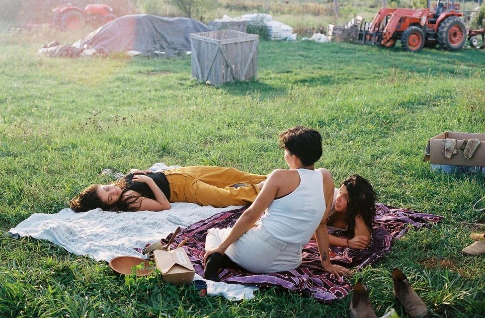 Three women relax on a blanket in a grassy field, enjoying the outdoors in warm sunlight. A tractor sits in the background.
