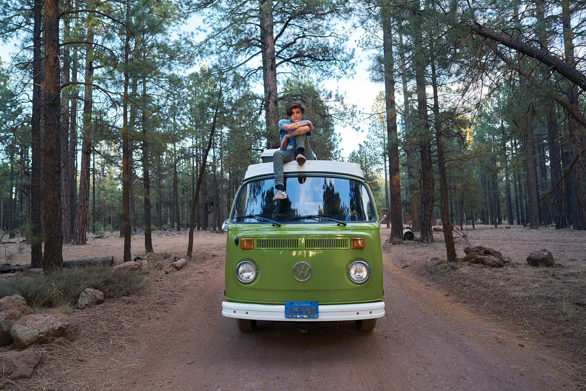 Young man sits on top of a green Volkswagen camper van, parked on a dirt road in a forest with tall trees on a sunny day.