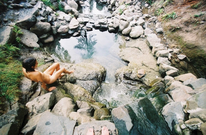 Woman seated on rocks beside a natural stone pool and waterfall, captured from a high-angle perspective.