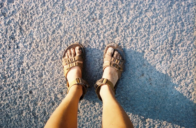 Overhead shot of muddy feet wearing brown buckled sandals standing on rough asphalt pavement.