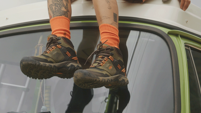 Close-up on hiking boots worn with orange socks, person sitting on top of a vintage vehicle, showcasing outdoor footwear.