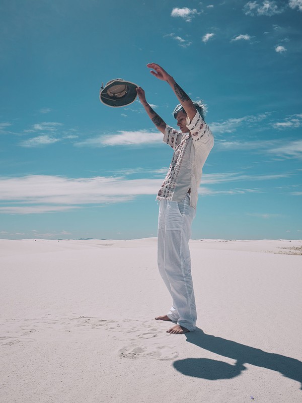 Man stands barefoot in the white sand of a desert, holding a hat above his head under a partly cloudy blue sky on a sunny day.