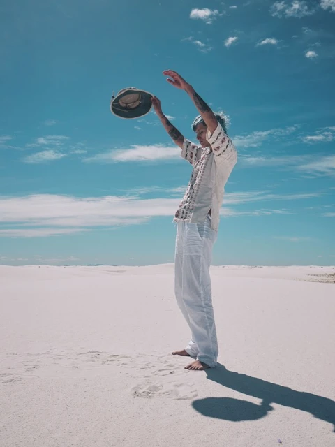 Man stands barefoot in the white sand of a desert, holding a hat above his head under a partly cloudy blue sky on a sunny day.