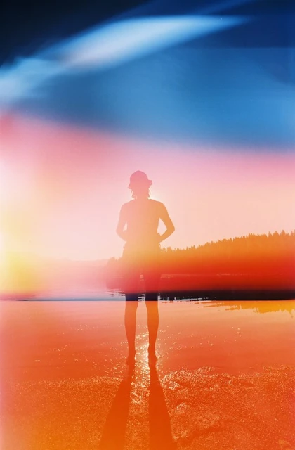 Silhouette of a person standing in shallow water on a beach during sunset with trees and water in the background and color film effects.