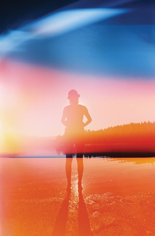 Person standing on a beach at sunset with lens flare. Silhouette of a person wearing a hat stands on the sand near the water's edge.