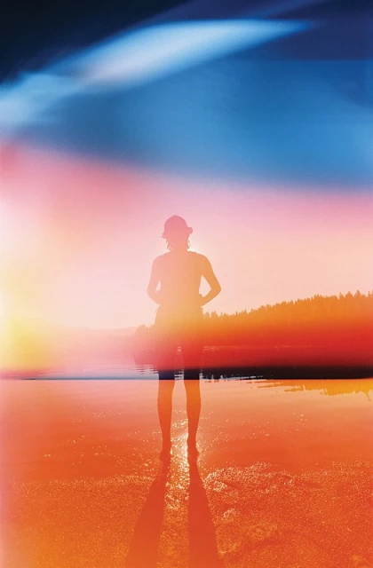 Person standing on a beach at sunset with lens flare. Silhouette of a person wearing a hat stands on the sand near the water's edge.