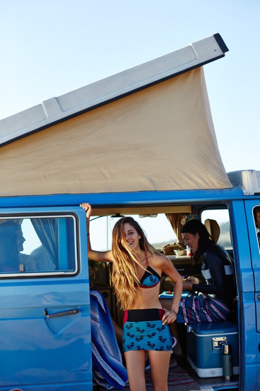 A woman in a bikini stands by a blue camper van on a sunny day, with another woman visible inside the vehicle.