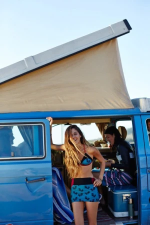 A woman in a bikini stands by a blue camper van on a sunny day, with another woman visible inside the vehicle.