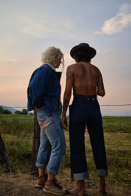 Two people standing in a rural landscape at dusk, wearing denim jackets and jeans, looking out over a field with a barbed wire fence.