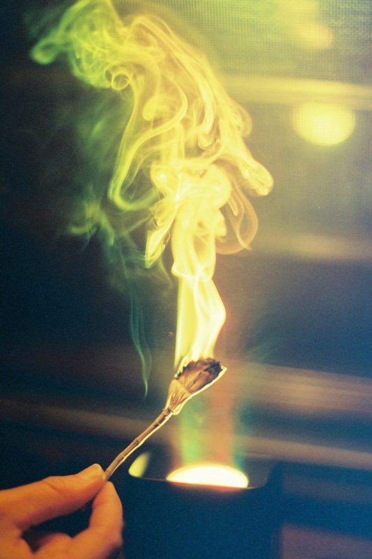 Hand holding burning botanical material with smoke rising, lit by a bright light in a dark environment.