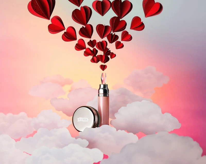 La Mer skincare products on clouds with red paper hearts floating above against a gradient pink and yellow background, beauty aesthetic.