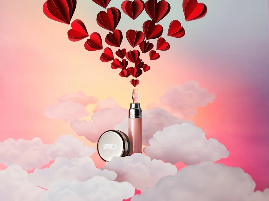 Cosmetic products, La Mer face powder and a lip gloss, are displayed on a cloud-like surface with red paper hearts against a pink-to-purple sky.