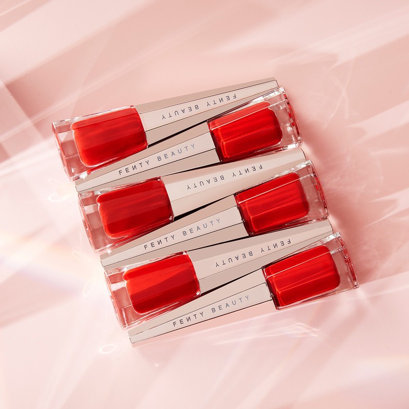 Stack of four Fenty Beauty lip gloss tubes featuring a clear rectangular design and vibrant red product against a pink gradient background.