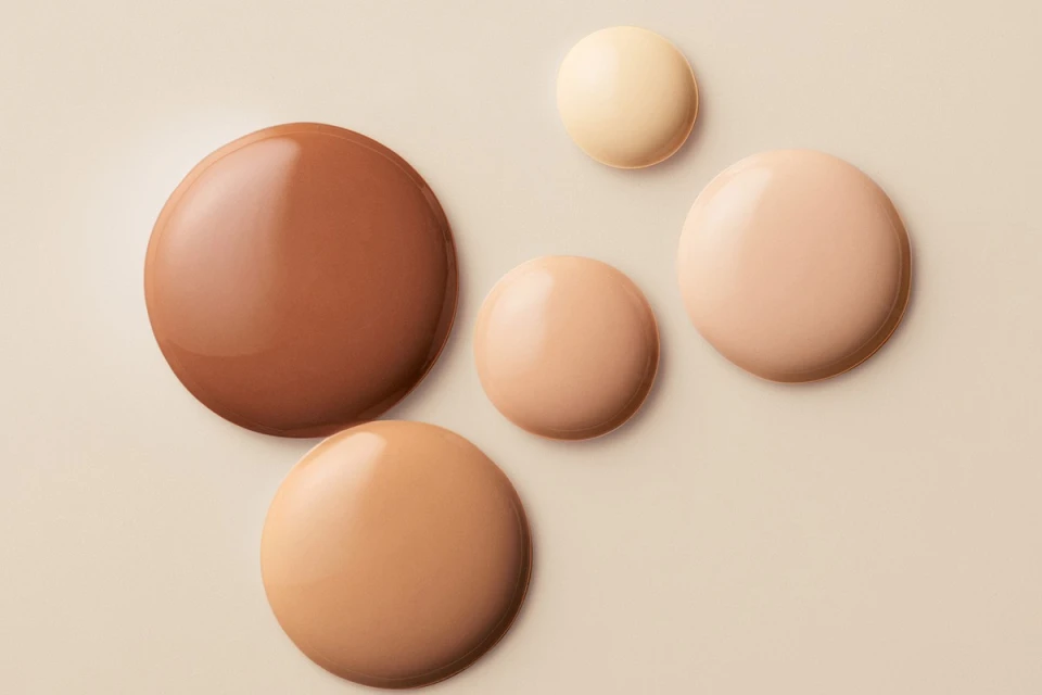 Five drops of different shades of foundation arranged on a light beige background, ranging from light to dark tone. Beauty concept.