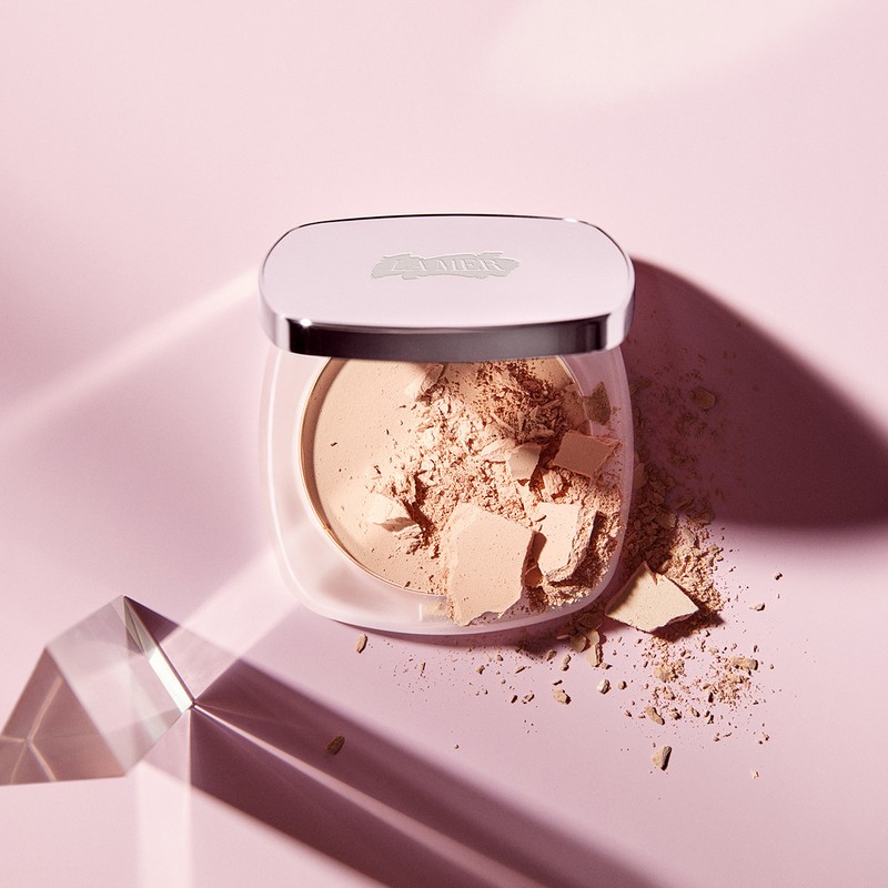 Close-up of a translucent powder compact filled with loose powder, casting shadows on a light pink background with a prism.