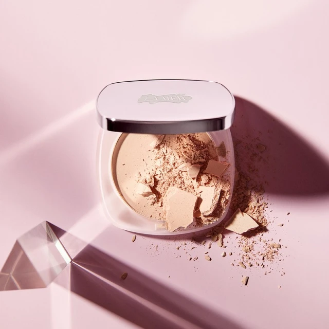 Close-up of a translucent powder compact filled with loose powder, casting shadows on a light pink background with a prism.