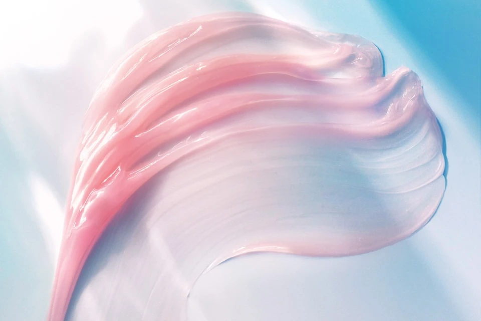 Close-up of a pink and white creamy cosmetic product swatch on a light blue background, highlighting its smooth texture and pearlescent sheen.