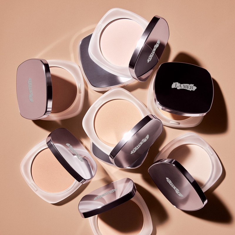 Overhead studio shot showcases a collection of La Mer pressed powders with reflective cases and product variations against a tan backdrop.