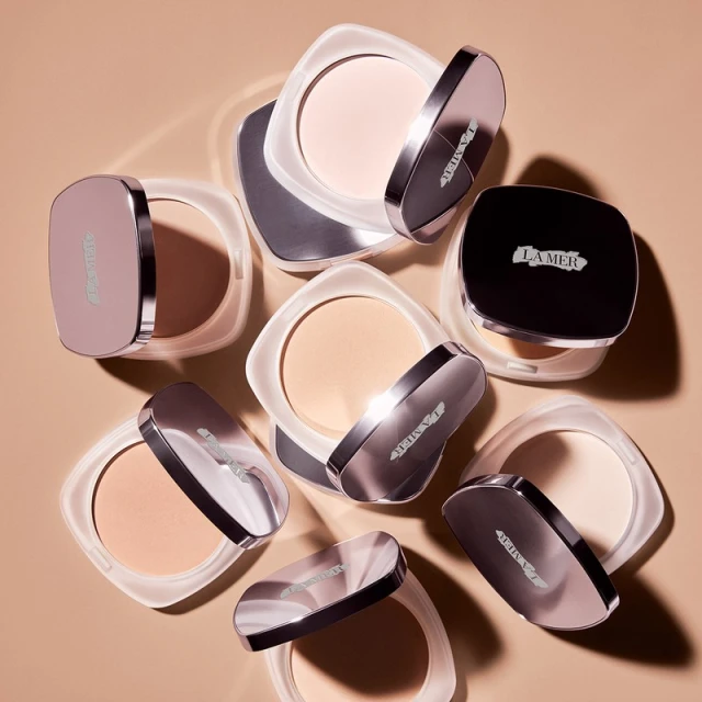 Overhead studio shot showcases a collection of La Mer pressed powders with reflective cases and product variations against a tan backdrop.