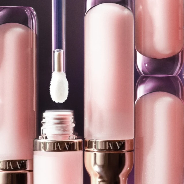 Close-up of pink lip gloss bottles with applicator wand and gold accents on a dark background for a beauty advertisement.