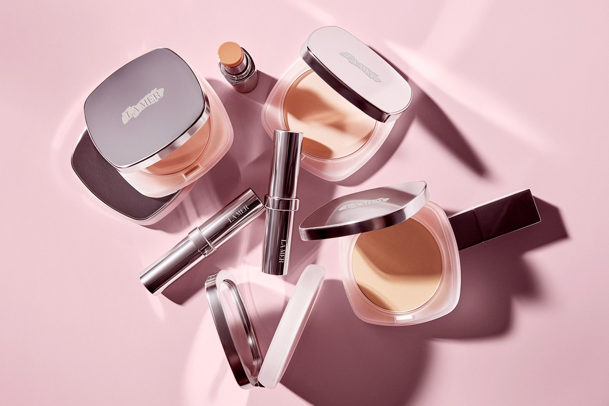 La Mer makeup products including pressed powder and concealer are arranged on a pink surface with light and shadow creating visual interest.