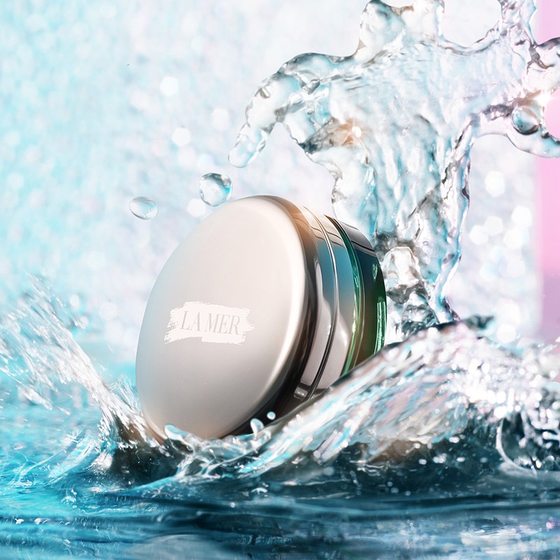 La Mer skincare product in a white jar, surrounded by water splashing against a blue and pink background for a beauty advertisement.