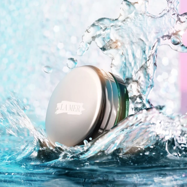 La Mer skincare product in a white jar, surrounded by water splashing against a blue and pink background for a beauty advertisement.