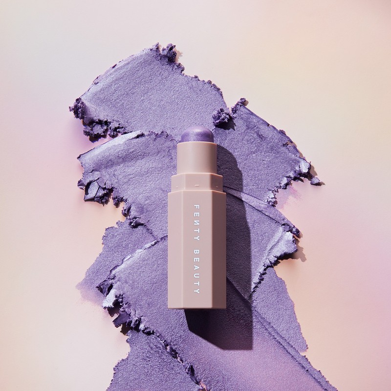 Fenty Beauty Match Stix Cream Shimmer with a swipe of shimmery purple eye shadow on a gradient peach to lavender background.