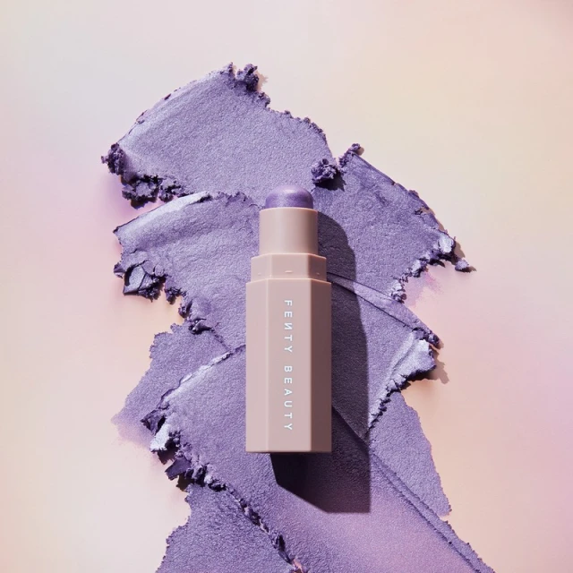 Fenty Beauty Match Stix Cream Shimmer with a swipe of shimmery purple eye shadow on a gradient peach to lavender background.