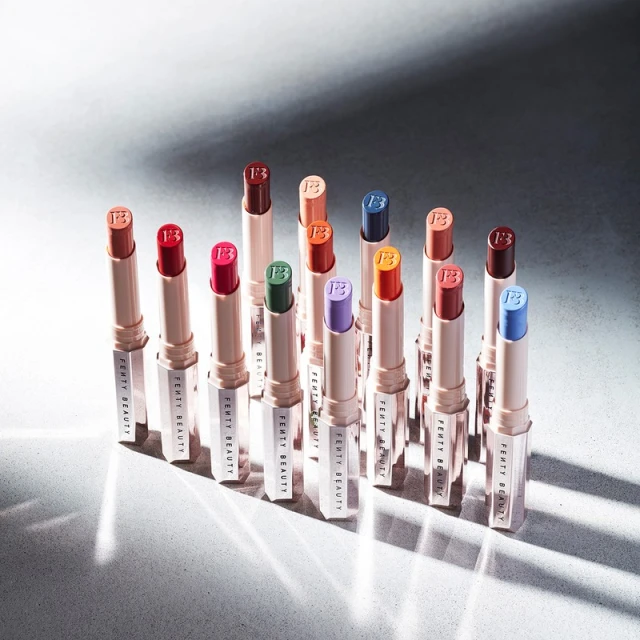 Fenty Beauty lipsticks in multiple colors, arranged in a group on a gray surface with studio lighting, displaying makeup products.