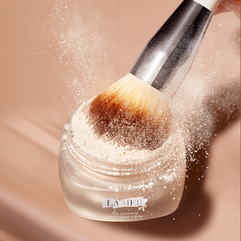 Close-up shot of a La Mer setting powder jar with a large brush, powder dusting in air, soft focus, and neutral beige color palette.