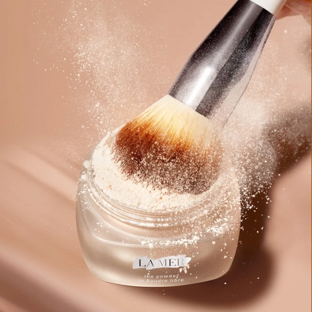 Close-up shot of a La Mer setting powder jar with a large brush, powder dusting in air, soft focus, and neutral beige color palette.